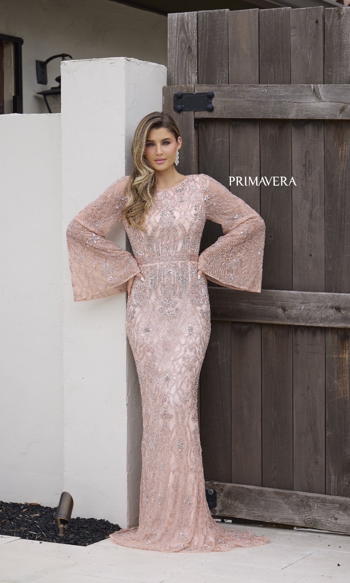 A woman wears the Primavera 12187, a fitted, long formal dress in light pink with beaded sequins, long bell sleeves, and a subtle pattern as she stands against a wooden gate.